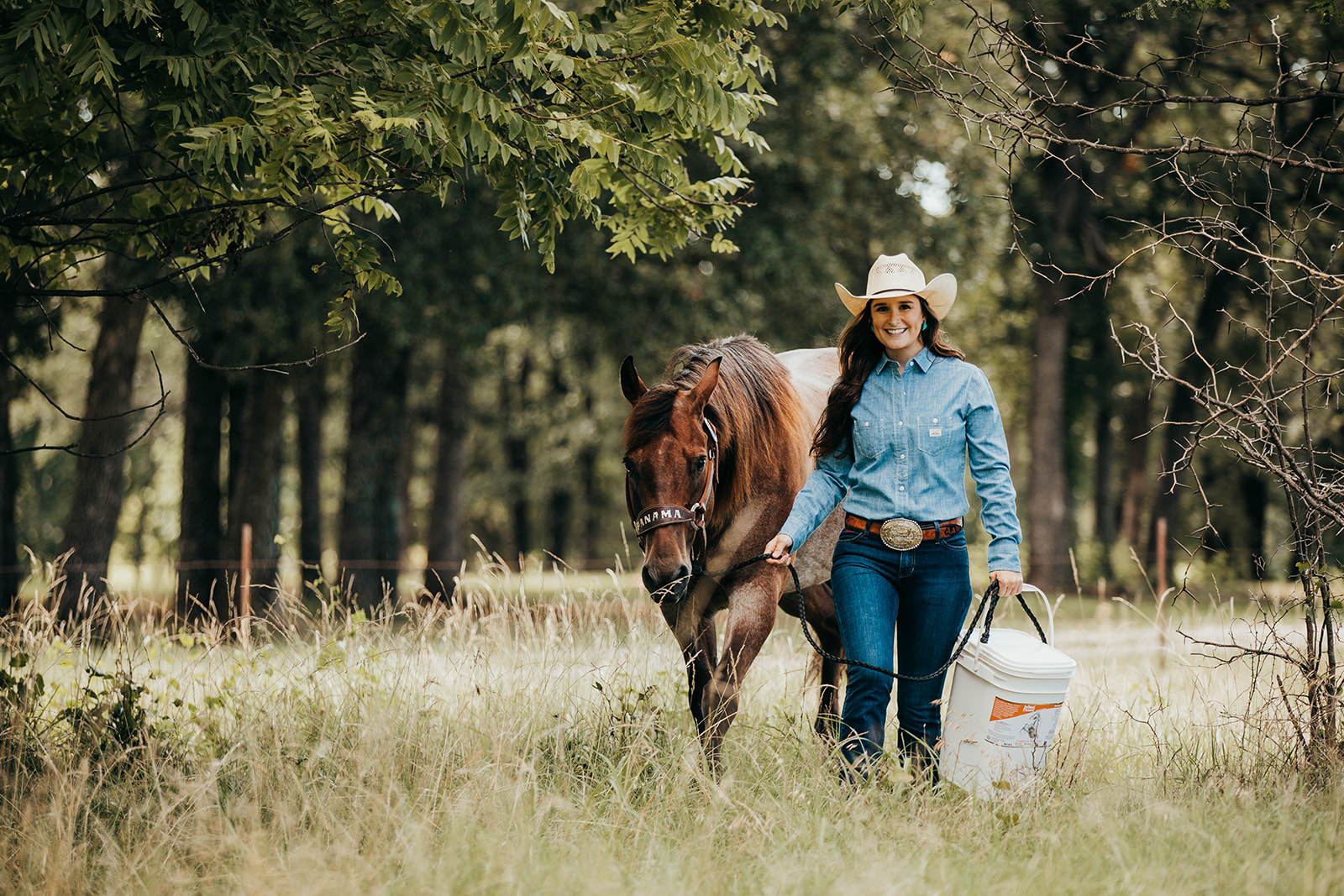 woman-in-blue-shirt-and-white-cowboy-hat-walking-her-horse-while-carrying-a-bucket-of-fdd